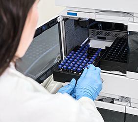 Scientist in a white lab coat putting vial with a sample into autosampler of HPLC system. High performance liquid chromatography at chemical laboratory.